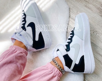 May include: White and black high-top sneakers with a black swoosh logo and a white sole. The sneakers are worn by a person wearing pink sweatpants and a silver chain bracelet.