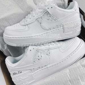 May include: White leather low-top sneakers with a pearl-embellished design. The sneakers have a white midsole and outsole, and the text "AIR" is printed on the side of the shoe. The text "Mrs. Tia" is written on the side of the shoe.
