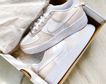May include: A pair of white and light beige sneakers with a white swoosh logo and the word "AIR" on the side. The sneakers are in a shoebox.