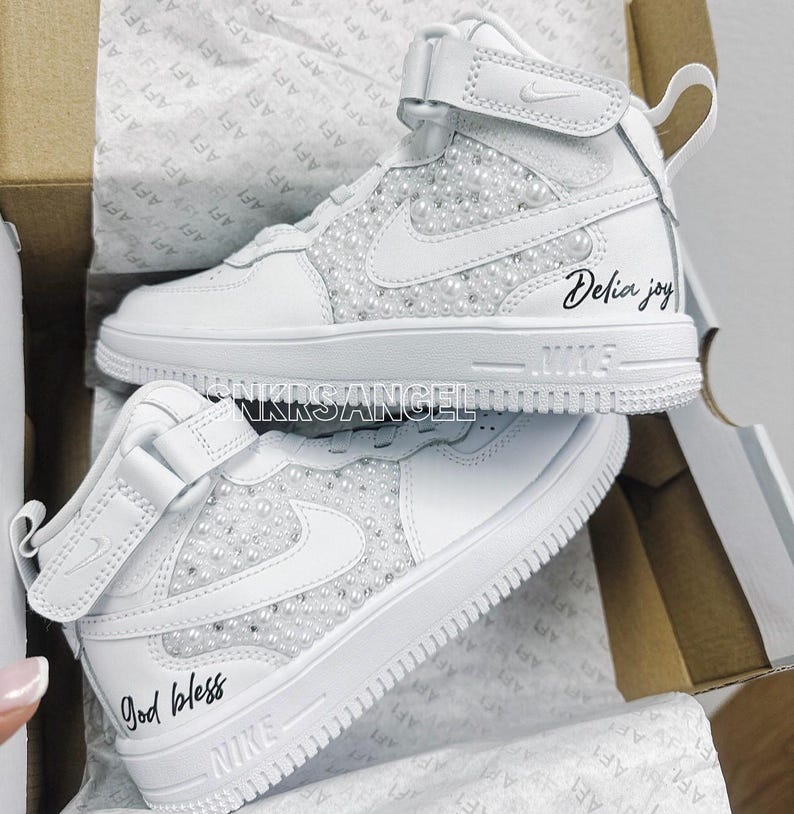 K&ouml;nnte beinhalten: Zwei wei&szlig;e High-Top-Sneaker mit Perlenbesatz. Ein Schuh hat den Text "God bless" darauf geschrieben, der andere den Text "Delia joy".