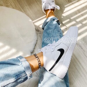 May include: White high-top trainers with a black Nike swoosh and a silver chain ankle bracelet. The trainers are worn by a person wearing blue jeans.