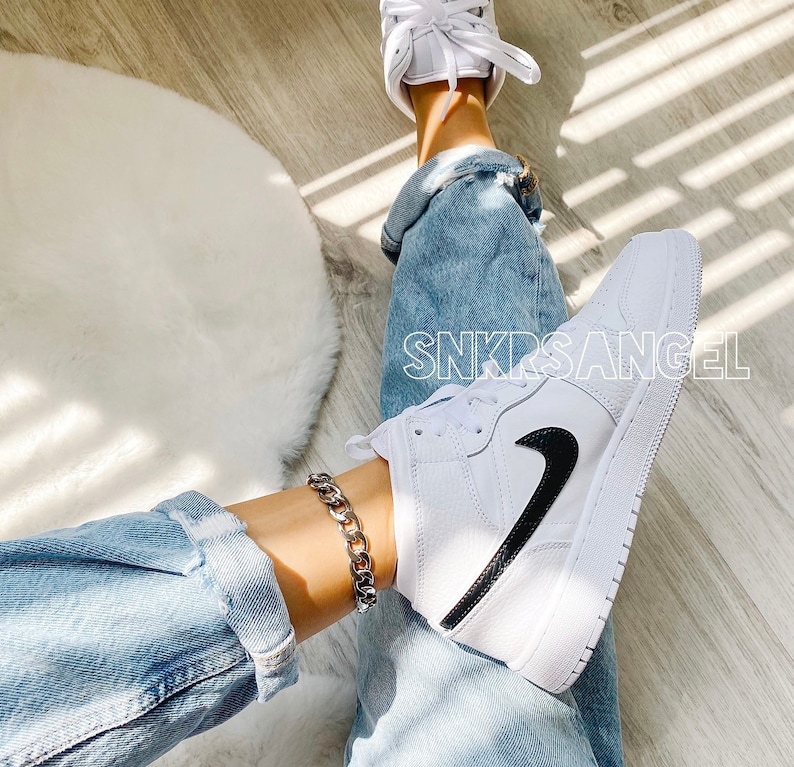 May include: White high-top trainers with a black Nike swoosh and a silver chain ankle bracelet. The trainers are worn by a person wearing blue jeans.