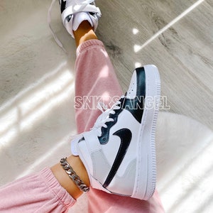 May include: White high-top trainers with a black swoosh and black trim. The trainers are worn with pink sweatpants and a silver chain ankle bracelet.
