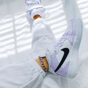 Op de afbeelding: Witte high-top sneakers met een paars en wit kleurblok ontwerp. De sneakers hebben een zwart swoosh logo en een dikke witte zool. De sneakers worden gedragen met een witte joggingbroek en een zilveren ketting enkelband.