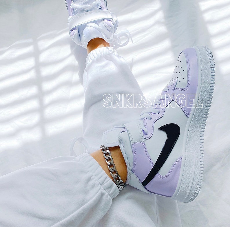 Op de afbeelding: Witte high-top sneakers met een paars en wit kleurblok ontwerp. De sneakers hebben een zwart swoosh logo en een dikke witte zool. De sneakers worden gedragen met een witte joggingbroek en een zilveren ketting enkelband.