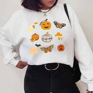 Może przedstawiać: Biała bluza z nadrukiem o tematyce Halloween z motywem dyni, motyli, ducha i grzyba. Na bluzie widnieje napis "Boo!" i "Little Pumpkins".