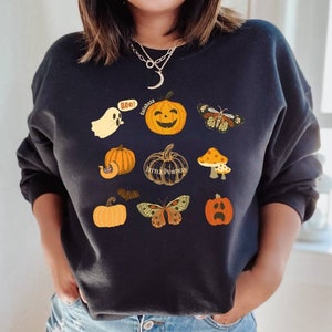 Może przedstawiać: Czarny sweter z motywem Halloween z dyniami, duchem, nietoperzem, motylami i grzybami. Tekst na swetrze brzmi "Boo!" i "Little Pumpkins".