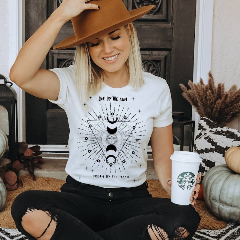 May include: White t-shirt with a black and white graphic design featuring a sun and moon with the text "Live by the sun, Dream by the moon".