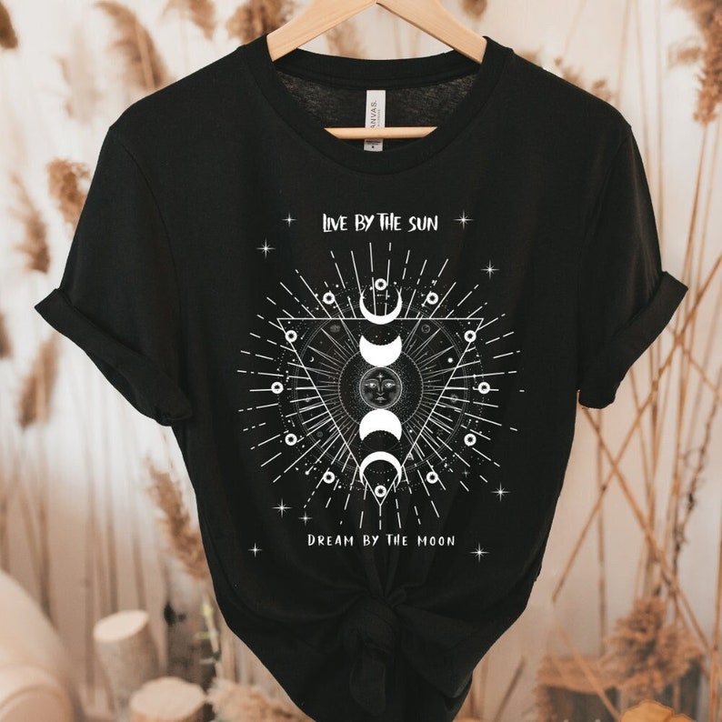 May include: Black t-shirt with a white graphic design featuring a sun and moon with the text "Live by the sun, Dream by the moon".
