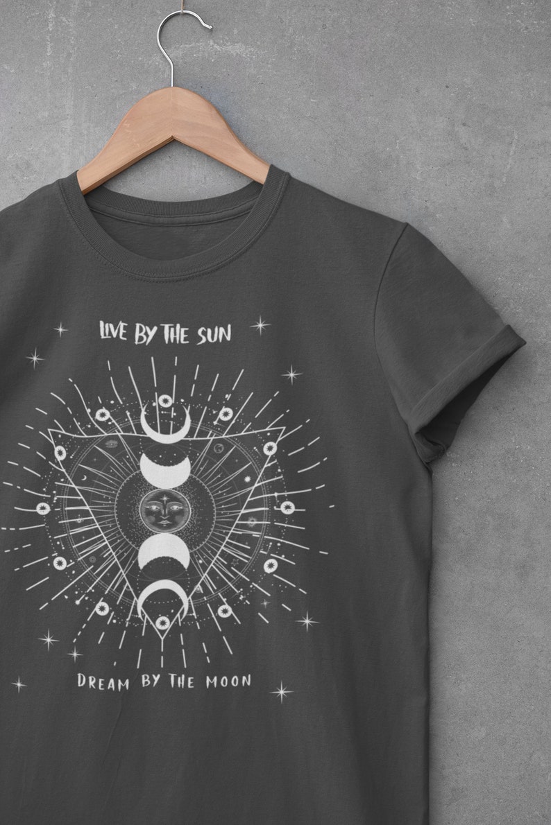 May include: A dark gray t-shirt with a white graphic design featuring a sun and moon with the text "Live by the sun, Dream by the moon".