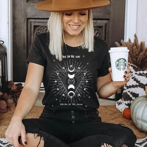 May include: Black t-shirt with a white graphic design featuring a sun and moon with the text "Live by the sun, Dream by the moon".