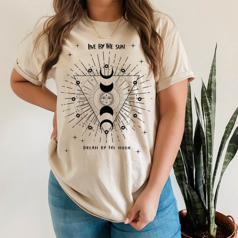 May include: A beige t-shirt with a black graphic design featuring a sun and moon with the text "Live by the sun, Dream by the moon".