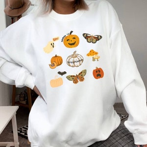 Może przedstawiać: Biała bluza z nadrukiem o tematyce Halloween z dyniami, duchem, nietoperzem, motylami i grzybami. Zawiera r&oacute;wnież tekst "Boo!" i "Calabaza".
