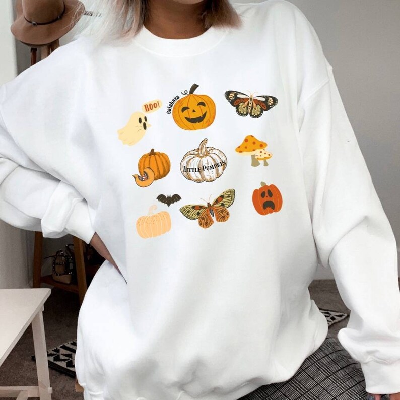 Może przedstawiać: Biała bluza z nadrukiem o tematyce Halloween z dyniami, duchem, nietoperzem, motylami i grzybami. Zawiera r&oacute;wnież tekst "Boo!" i "Calabaza".