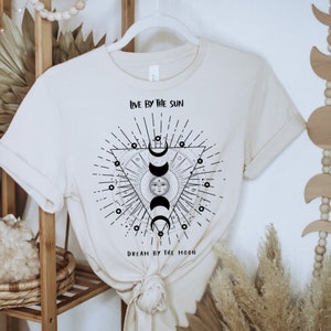 May include: A beige t-shirt with a black and white graphic design of a sun and moon in a triangle with the text "Live by the sun, Dream by the moon".