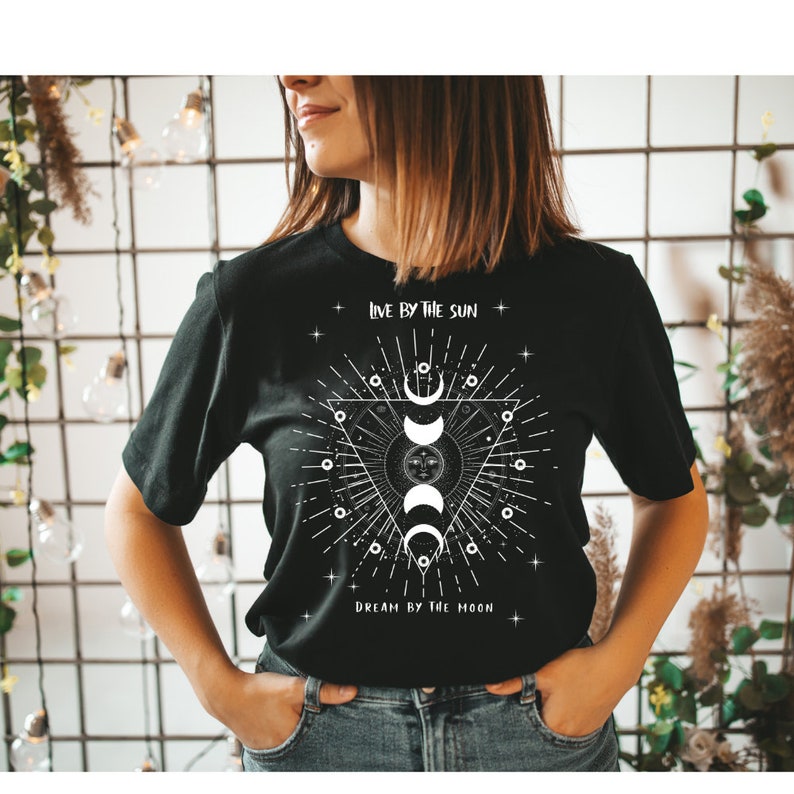 May include: Black t-shirt with a white graphic design featuring a sun and moon with the text "Live by the sun, Dream by the moon."