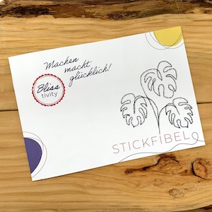 May include: A white card with black line drawings of monstera leaves and the text "STICKFIBEL" in red. The card also has a purple circle and a yellow circle. The text "Machen macht gl&uuml;cklich!" is written in black cursive. A red circle with the text "Bliss tivity" is in the top left corner.