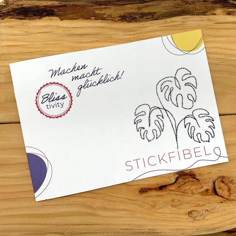 May include: A white card with black line drawings of monstera leaves and the text "STICKFIBEL" in red. The card also has a purple circle and a yellow circle. The text "Machen macht gl&uuml;cklich!" is written in black cursive. A red circle with the text "Bliss tivity" is in the top left corner.