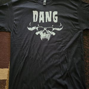 May include: A black short-sleeve t-shirt featuring the word DANG printed in white capital letters above a white graphic illustration of a horned skull with sharp teeth. The shirt has a crew neckline and a standard fit with a visible brand label.