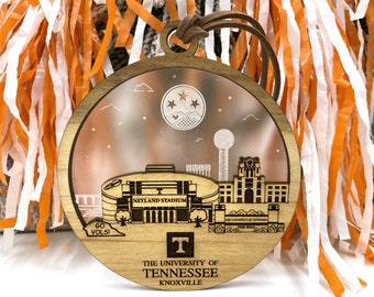 May include: A wooden ornament shaped like a circle with a depiction of Neyland Stadium, the University of Tennessee, and the city of Knoxville. The ornament is brown with a clear, frosted center. The text "The University of Tennessee Knoxville" is printed on the bottom of the ornament.