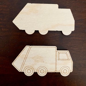 May include: Two unfinished wooden cutouts of garbage trucks. One is a solid shape, and the other has etched details including wheels, a cab, and a loading area. The trucks are light brown and sit on a dark brown surface.