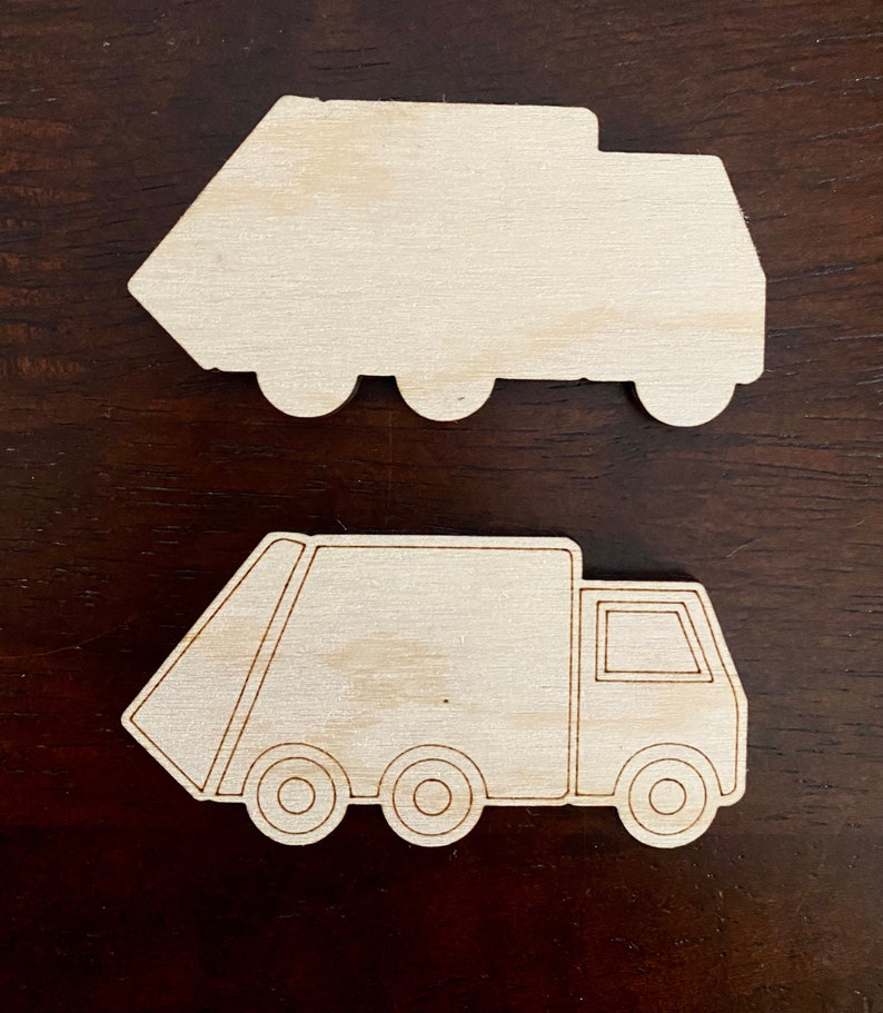 May include: Two unfinished wooden cutouts of garbage trucks. One is a solid shape, and the other has etched details including wheels, a cab, and a loading area. The trucks are light brown and sit on a dark brown surface.