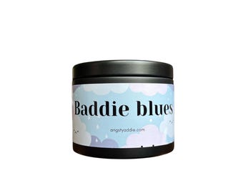 May include: A round, black container with a label featuring the text "Baddie blues" in a stylized font. The label has a light blue background with white clouds and the website address angstyaddie.com.