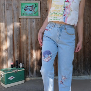 Peut inclure: Jean bleu clair avec des détails de patchwork, dont un motif circulaire et des étoiles. Une enseigne Pepsi-Cola vintage est accrochée à un mur en bois. Une glacière verte et des bouteilles sont également visibles.