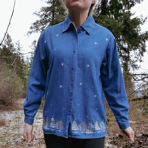 May include: A blue denim button-down shirt with long sleeves. The shirt features embroidered white snowflakes and a forest scene along the bottom hem. The shirt has a collared neckline and is buttoned up.