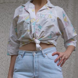 Peut inclure: Une chemise blanche à boutons avec des manches trois-quarts et un imprimé abstrait pastel jaune, rose et bleu sarcelle, nouée à la taille. La chemise est associée à un jean en denim bleu clair délavé, taille haute, avec des boutons en métal.
