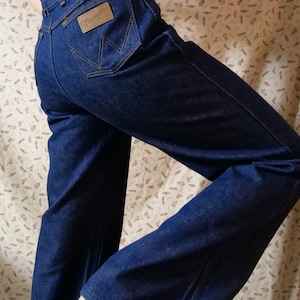 May include: Dark blue denim Wrangler jeans with a high waist and flared leg. The jeans feature a brown leather patch with the Wrangler logo on the back pocket. The jeans are worn with a tan top and espadrille shoes.