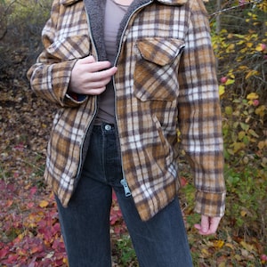 May include: A brown and white plaid jacket with a grey fleece lining and a silver zip. The jacket has two front pockets and is worn with dark grey jeans. The overall style is vintage and casual.