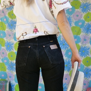 Peut inclure: Un jean Wrangler noir avec un patch en cuir sur la poche arrière. Un haut blanc brodé de motifs floraux roses est visible. Un chapeau de paille avec un ruban à pois noirs est tenu à la main.