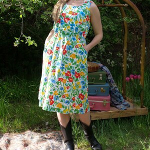 Peut inclure: Une robe sans manches arrivant au genou, dotée d'une base blanche avec un motif floral vif en rouge, jaune et bleu. La robe présente une encolure ronde avec un fin liseré rouge, une taille cintrée et des poches latérales, portée avec des bottes.