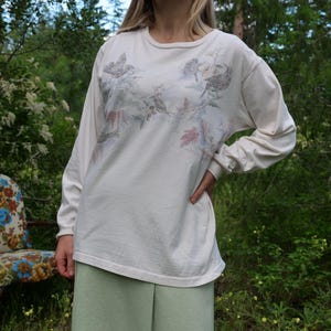 May include: A white long-sleeved shirt with a floral print featuring birds and leaves. The shirt has a crew neck and is made of a soft, comfortable fabric.