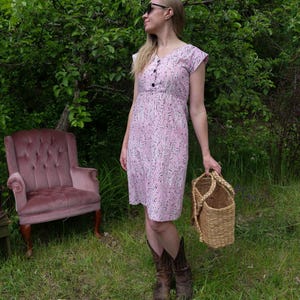 Peut inclure: Robe rose clair avec un motif abstrait noir et des boutons noirs. La robe est aux genoux avec des manches courtes. Un panier en osier est tenu à la main. Un fauteuil rose vintage est en arrière-plan.
