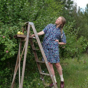 May include: A person in a floral print outfit stands on a wooden orchard ladder, reaching towards an apple tree. The outfit is a matching set of a short-sleeved shirt and skirt. Apples rest on a small shelf on the ladder.