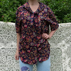 May include: A short-sleeved shirt with a dark navy base and a floral design in red, green, and orange. The shirt features a collar and a tie detail at the neckline. The model is wearing light blue jeans.