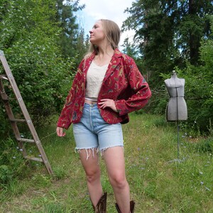 May include: A woman wearing a red floral blazer, light blue denim shorts, and brown cowboy boots. The blazer has a colorful floral pattern. A white crop top is visible underneath. A wooden ladder and a dress form are in the background.