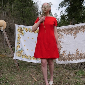 May include: A woman wearing a red dress stands in a grassy area with two floral tablecloths hanging behind her. The woman is holding a bouquet of white flowers in front of her face.