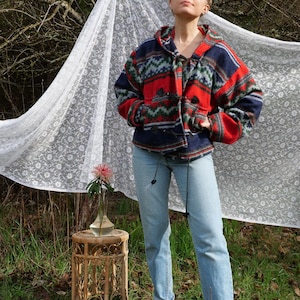 May include: A hooded fleece jacket in a red, blue, and green geometric design. The jacket features a drawstring and two front pockets. The person is wearing light blue jeans. A small wicker table holds a vase with a pink flower.