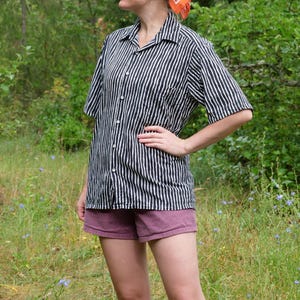 Peut inclure: Une chemise à manches courtes à rayures verticales noires et blanches. Elle est boutonnée et portée avec un short mauve. L'ensemble est porté en extérieur, dans un cadre naturel avec de la verdure et des fleurs sauvages.