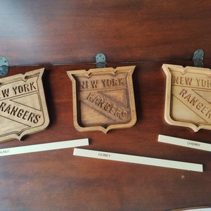 Puede incluir: Tres logotipos de los New York Rangers de madera, cada uno con una mancha de madera diferente. Los logotipos est&aacute;n sobre una superficie de madera con etiquetas para "Nogal especial", "Miel" y "Cereza".