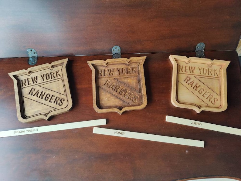Puede incluir: Tres logotipos de los New York Rangers de madera, cada uno con una mancha de madera diferente. Los logotipos est&aacute;n sobre una superficie de madera con etiquetas para "Nogal especial", "Miel" y "Cereza".