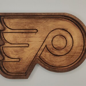 May include: A wooden wall decor in the shape of the Philadelphia Flyers logo. The logo is brown and features a stylized flying 'P' with a wing.