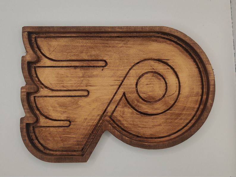 May include: A wooden wall decor in the shape of the Philadelphia Flyers logo. The logo is brown and features a stylized flying 'P' with a wing.