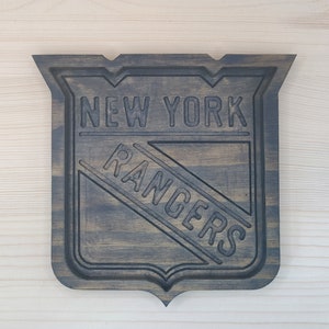 Puede incluir: Un letrero de madera marr&oacute;n oscuro con el logotipo de los New York Rangers. El logotipo es una letra "R" blanca dentro de un tri&aacute;ngulo azul con un contorno blanco. Las palabras "New York Rangers" est&aacute;n talladas en la madera.