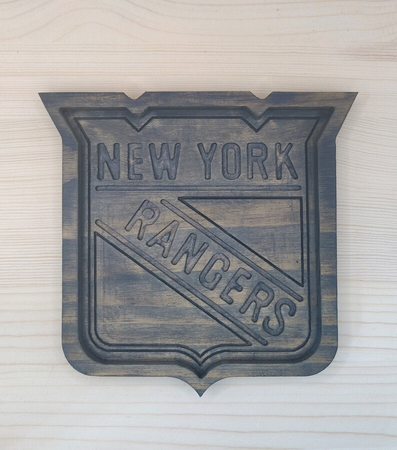Puede incluir: Un letrero de madera marr&oacute;n oscuro con el logotipo de los New York Rangers. El logotipo es una letra "R" blanca dentro de un tri&aacute;ngulo azul con un contorno blanco. Las palabras "New York Rangers" est&aacute;n talladas en la madera.