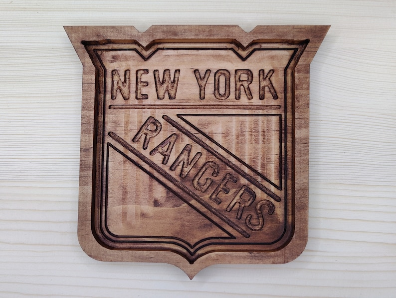 Puede incluir: Un letrero de madera con el logotipo de los New York Rangers. El logotipo es un escudo con las palabras "New York Rangers" en color marr&oacute;n oscuro.