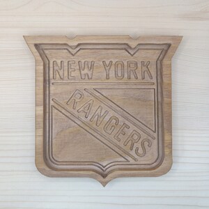 Puede incluir: Un letrero de madera con el logotipo de los New York Rangers. El logotipo est&aacute; tallado en la madera e incluye el nombre del equipo y una franja diagonal.
